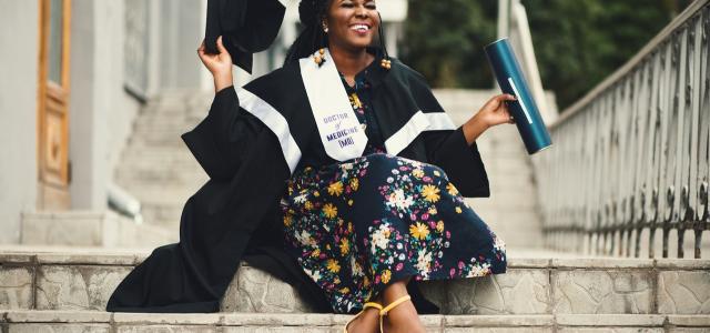 Woman in grad cap and gown sitting on stairs and smiling. She's wearing a ribbon that says Doctor of Medicine